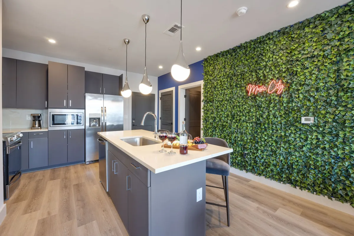 A lively kitchen scene featuring a prominent kitchen island, a neon sign, a lush green accent wall, bar stools, and wine glasses. The arrangement encourages both cooking and socializing, with the bar area clearly integrated into the open kitchen space.
