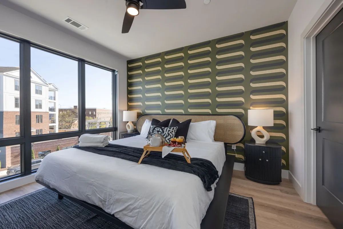 A serene primary bedroom featuring a king bed, an artful feature wall, and large windows that let in streams of natural light. Soft lighting fixtures add a cozy, hotel-inspired ambiance, while the overall layout feels spacious and inviting.