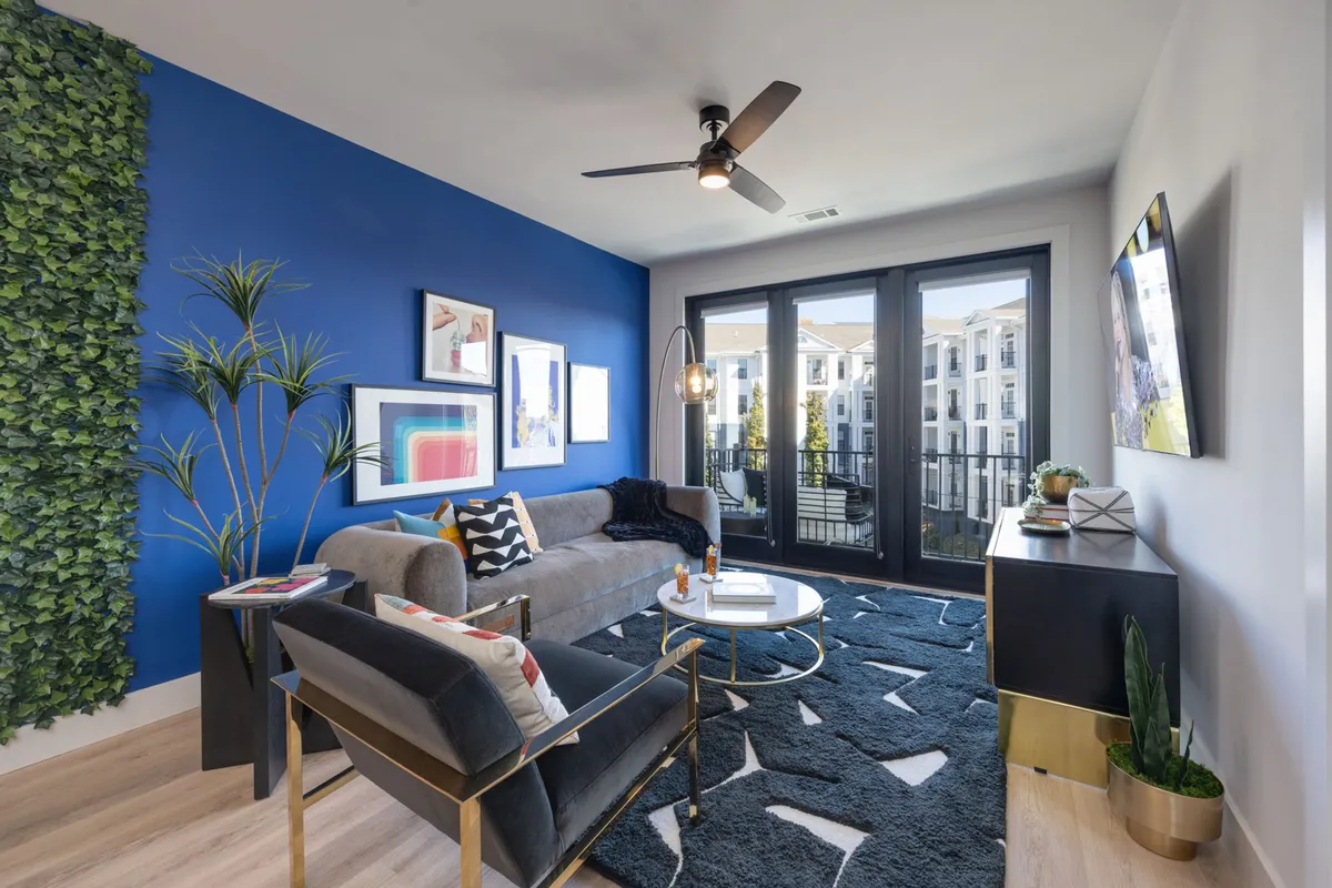 The image showcases a modern living room with a striking green accent wall, bold artwork, a plush sofa arrangement, and sliding glass doors that open to a balcony with sunrise views. The setting exudes both contemporary style and cozy comfort, inviting relaxation and social gatherings.
