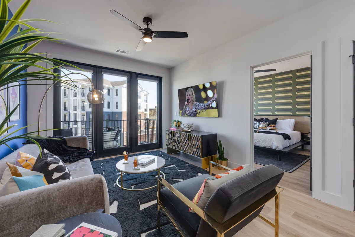 A contemporary living room featuring a comfortable sofa, coffee table, a large TV, and a balcony with outdoor seating. The room is brightly lit by natural sunlight streaming through large windows, and a potted plant adds freshness to the space. The open balcony offers city views, enhancing the indoor-outdoor connection.