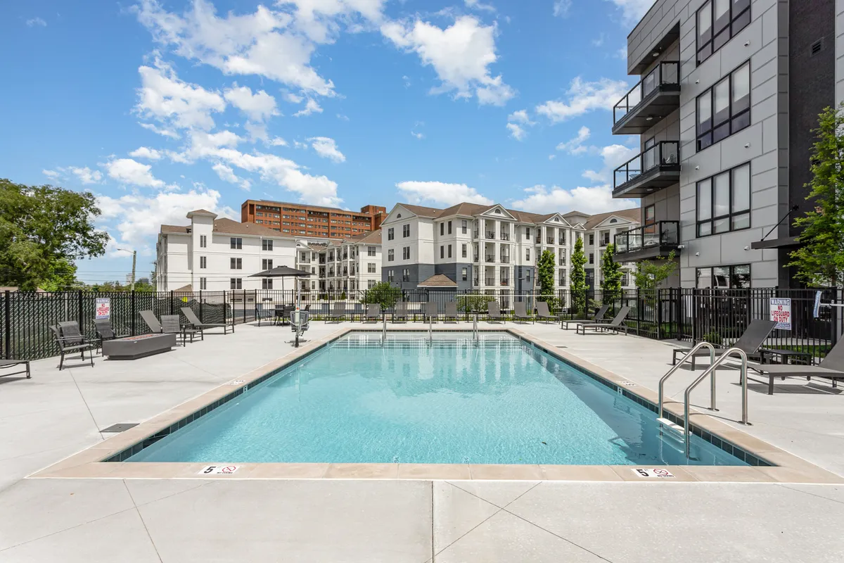 A view of the sparkling swimming pool with lounge chairs arranged around the deck, framed by modern apartment buildings and an open sky. The image evokes leisure, community, and an urban oasis vibe.