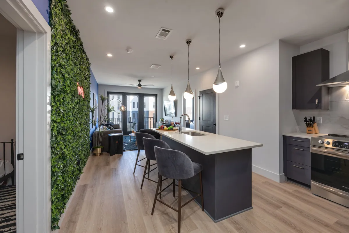 A spacious kitchen and dining zone with a kitchen island, bar stools, pendant lighting, a feature green wall, and an open balcony with outdoor seating. The space is stylish and airy, seamlessly connecting indoor dining with outdoor relaxation.