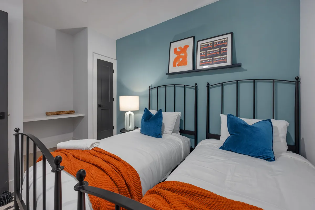 A cheerful second bedroom with two twin beds accented by blue pillows and a bright orange throw blanket. Framed artwork and a modern lamp add personality, making it ideal for families or friends seeking a welcoming, restful space.