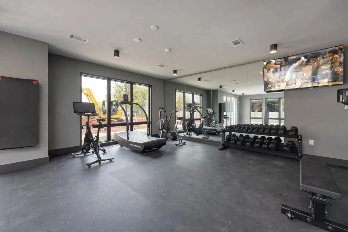 A bright, modern fitness center equipped with treadmills, a weights rack, an exercise bike, and a mirrored wall. Large windows bring in natural light, creating an energetic environment for personal and group workouts.