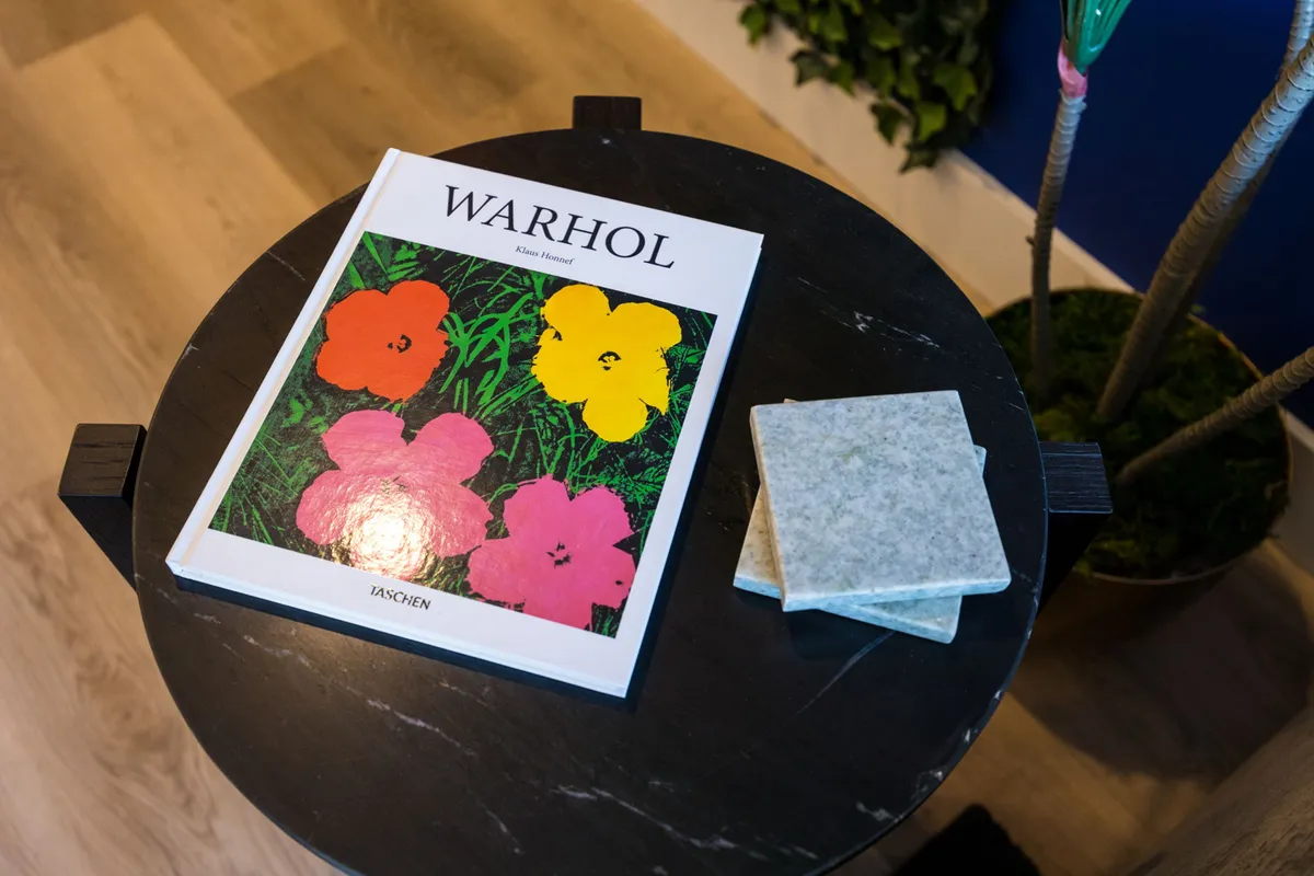 A stylish tabletop featuring a vibrant 'WARHOL' art book, colorful flower illustrations, and decorative coasters, evoking a lively and creative atmosphere for socializing, relaxing, or starting an engaging conversation.