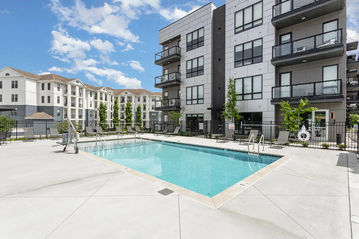 A sparkling swimming pool surrounded by landscaped greenery, with the apartment building's balconies overlooking the scene. The pool deck features lounge chairs and is bathed in warm sunlight, creating an inviting resort-style atmosphere ideal for relaxation and socializing.